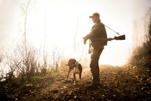 Hunter with rifle standing on gravel road and holding dog on a leash. Bright light is in the background.